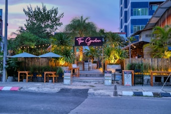 A vibrant outdoor seating area of a restaurant called 'The Garden' is visible. The entrance is adorned with lush greenery, including palm trees and various plants. Tables and chairs are set under patio umbrellas, with string lights enhancing the inviting atmosphere. The restaurant has a modern facade, and a neon sign is prominently displayed above the entrance. An adjacent building with multiple floors is partially visible in the background.