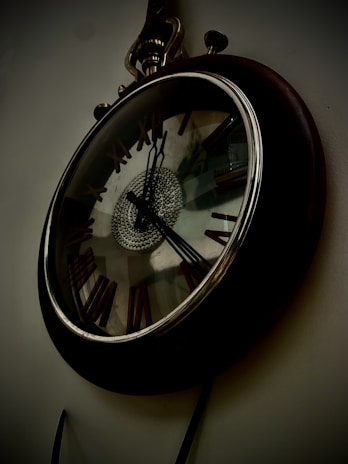A vintage wall clock with Roman numerals and ornate metal detailing. The clock has a reflective glass surface with a circular pattern in the center. Its hands are set to approximately 5:10.