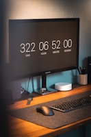 A desk with a computer monitor displaying a countdown timer. The setup includes a keyboard and mouse on a dark mat, and a small round device beside the monitor. There's also a smartwatch dock on the desk. The workspace is tidy and organized, set against a light-colored wall.