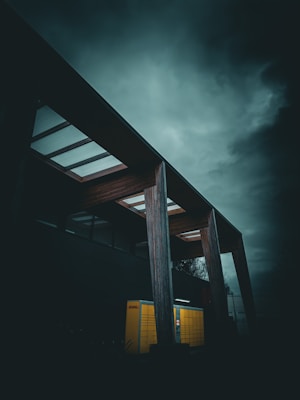 A dark, moody scene features a modern structure with large wooden beams and a translucent roof. Beneath, a yellow parcel locker stands out vividly against the gloomy sky, creating a stark contrast with the surrounding shadows.