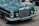 A vintage vehicle with a prominent grille and classic circular headlights is parked on the street. The car features a shiny chrome finish and intricate detailing, characteristic of older luxury automobiles. The reflection of trees is visible on the dark, polished surface of the car.