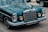 A vintage vehicle with a prominent grille and classic circular headlights is parked on the street. The car features a shiny chrome finish and intricate detailing, characteristic of older luxury automobiles. The reflection of trees is visible on the dark, polished surface of the car.