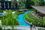 A serene outdoor setting featuring a winding blue swimming pool surrounded by lush greenery and hedges. Nearby, a wooden-roofed pavilion with outdoor dining tables offers a place to relax. In the foreground, three people walk along a pathway that bridges over the pool. Residential buildings with balconies enclose the area, and tall trees provide shade and add to the tranquil ambiance.