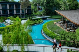 A serene outdoor setting featuring a winding blue swimming pool surrounded by lush greenery and hedges. Nearby, a wooden-roofed pavilion with outdoor dining tables offers a place to relax. In the foreground, three people walk along a pathway that bridges over the pool. Residential buildings with balconies enclose the area, and tall trees provide shade and add to the tranquil ambiance.
