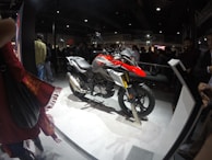 A motorcycle is displayed on a platform in a crowded indoor venue. The motorcycle features a combination of red, silver, and black colors, with the BMW logo visible. Several people are gathered around, observing the display. The environment is dimly lit, with the spotlight on the motorcycle, creating a contrast against the dark background.