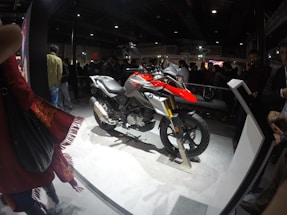 A motorcycle is displayed on a platform in a crowded indoor venue. The motorcycle features a combination of red, silver, and black colors, with the BMW logo visible. Several people are gathered around, observing the display. The environment is dimly lit, with the spotlight on the motorcycle, creating a contrast against the dark background.