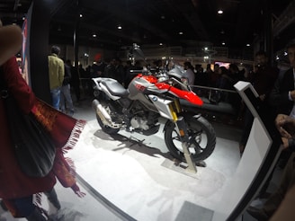 A motorcycle is displayed on a platform in a crowded indoor venue. The motorcycle features a combination of red, silver, and black colors, with the BMW logo visible. Several people are gathered around, observing the display. The environment is dimly lit, with the spotlight on the motorcycle, creating a contrast against the dark background.