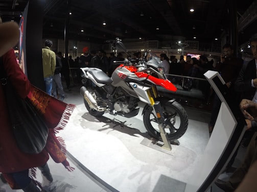 A motorcycle is displayed on a platform in a crowded indoor venue. The motorcycle features a combination of red, silver, and black colors, with the BMW logo visible. Several people are gathered around, observing the display. The environment is dimly lit, with the spotlight on the motorcycle, creating a contrast against the dark background.