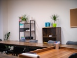 A modern office space with wooden tables arranged in a U-shape, black chairs, and open shelving. The shelves hold various office supplies, plants, and books. A corkboard hangs on a white wall near a green potted plant. Brightly colored mugs and a plant are placed on a shelf.