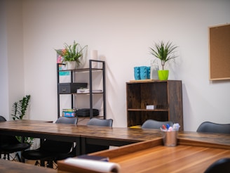 A modern office space with wooden tables arranged in a U-shape, black chairs, and open shelving. The shelves hold various office supplies, plants, and books. A corkboard hangs on a white wall near a green potted plant. Brightly colored mugs and a plant are placed on a shelf.
