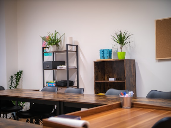 A modern office space with wooden tables arranged in a U-shape, black chairs, and open shelving. The shelves hold various office supplies, plants, and books. A corkboard hangs on a white wall near a green potted plant. Brightly colored mugs and a plant are placed on a shelf.