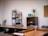 A modern office space with wooden tables arranged in a U-shape, black chairs, and open shelving. The shelves hold various office supplies, plants, and books. A corkboard hangs on a white wall near a green potted plant. Brightly colored mugs and a plant are placed on a shelf.
