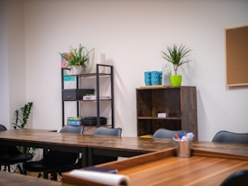 A modern office space with wooden tables arranged in a U-shape, black chairs, and open shelving. The shelves hold various office supplies, plants, and books. A corkboard hangs on a white wall near a green potted plant. Brightly colored mugs and a plant are placed on a shelf.