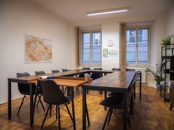 A well-organized meeting room with wooden floors and a large, L-shaped conference table surrounded by black chairs. Two large windows allow natural light into the space, with a map and colorful drawings on the walls. A clock is mounted above the radiators, and plants add a touch of greenery to the minimalistic decor. Shelving holds books and other items.