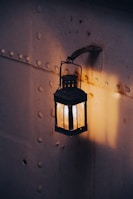 A vintage lantern hangs on a textured metal wall, softly illuminating its surroundings with a warm, golden light. The wall is riveted and shows signs of rust and wear, adding a touch of nostalgic ambiance.