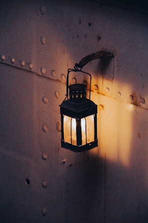 A vintage lantern hangs on a textured metal wall, softly illuminating its surroundings with a warm, golden light. The wall is riveted and shows signs of rust and wear, adding a touch of nostalgic ambiance.