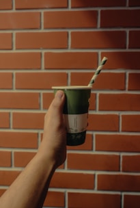 A hand holding a green disposable cup with a paper straw against a background of a red brick wall.