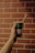 A hand holding a green disposable cup with a paper straw against a background of a red brick wall.