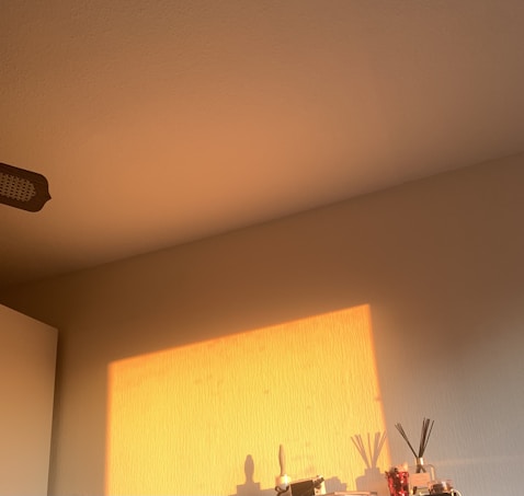 A soft, warm sunlight casts a rectangular pattern on a beige wall, creating gentle shadows. A shelf at the bottom holds various items, including a red-tinted diffuser with black reeds and several bottles or containers.