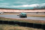 A green car is drifting around a curve on a racetrack with blurred motion, suggesting high speed. The surroundings include a grassy area and a barrier made of stacked tires. In the background, another vehicle is visible, and the sky is overcast.