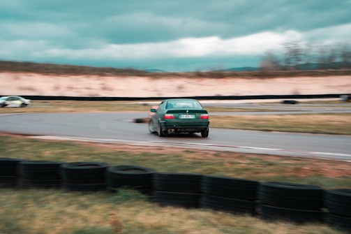 A green car is drifting around a curve on a racetrack with blurred motion, suggesting high speed. The surroundings include a grassy area and a barrier made of stacked tires. In the background, another vehicle is visible, and the sky is overcast.