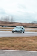 A silver car is drifting on a racetrack with motion blur indicating high speed. The background shows an overcast sky and a blurred landscape, suggesting a focus on the car's motion. Other features include worn-out asphalt and grassy areas.