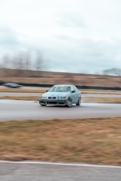 A silver car is drifting on a racetrack with motion blur indicating high speed. The background shows an overcast sky and a blurred landscape, suggesting a focus on the car's motion. Other features include worn-out asphalt and grassy areas.