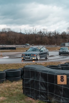 A dark-colored car with noticeable decals is drifting on a wet race track, surrounded by tire barriers. The background features leafless trees and a cloudy sky, creating a typical motorsport scene.