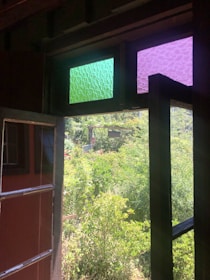 An open window with two upper panes featuring textured green and purple glass overlooks a lush garden filled with dense greenery and various plants capturing natural light.