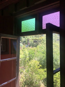 An open window with two upper panes featuring textured green and purple glass overlooks a lush garden filled with dense greenery and various plants capturing natural light.