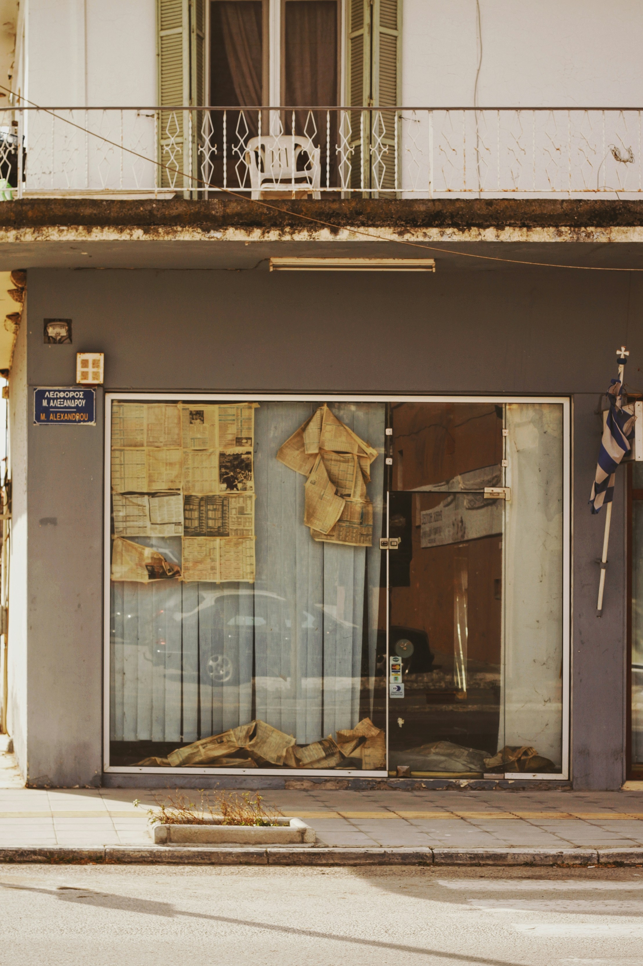 A storefront window covered with old newspapers and blinds is seen below a residential balcony with green wooden shutters and a white plastic chair. The facade shows signs of neglect, with an overgrown plant on the pavement and a weathered Greek flag beside the entrance.