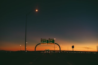 A highway scene during sunset with an overpass and road signs indicating directions to Van Horn and El Paso. The sky displays a gradient of colors from deep blue at the top to a warm orange near the horizon, with the silhouette of distant mountains. Streetlights and vehicles are visible, adding to the urban setting.