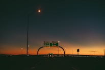 A highway scene during sunset with an overpass and road signs indicating directions to Van Horn and El Paso. The sky displays a gradient of colors from deep blue at the top to a warm orange near the horizon, with the silhouette of distant mountains. Streetlights and vehicles are visible, adding to the urban setting.