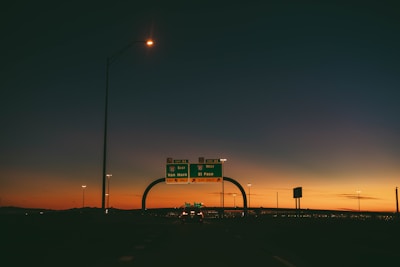 A highway scene during sunset with an overpass and road signs indicating directions to Van Horn and El Paso. The sky displays a gradient of colors from deep blue at the top to a warm orange near the horizon, with the silhouette of distant mountains. Streetlights and vehicles are visible, adding to the urban setting.