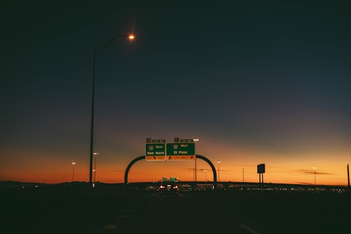 A highway scene during sunset with an overpass and road signs indicating directions to Van Horn and El Paso. The sky displays a gradient of colors from deep blue at the top to a warm orange near the horizon, with the silhouette of distant mountains. Streetlights and vehicles are visible, adding to the urban setting.