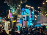 A vibrant, bustling outdoor nighttime scene featuring a lively cafe or entertainment venue with string lights and neon signs. People are seated at tables, socializing and enjoying the atmosphere beneath a large tree, surrounded by colorful signage that adds a modern and lively touch.