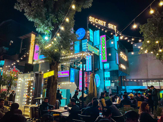 A vibrant, bustling outdoor nighttime scene featuring a lively cafe or entertainment venue with string lights and neon signs. People are seated at tables, socializing and enjoying the atmosphere beneath a large tree, surrounded by colorful signage that adds a modern and lively touch.