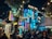A vibrant, bustling outdoor nighttime scene featuring a lively cafe or entertainment venue with string lights and neon signs. People are seated at tables, socializing and enjoying the atmosphere beneath a large tree, surrounded by colorful signage that adds a modern and lively touch.