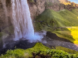 A powerful waterfall cascades down from a rocky cliff into a misty pool below, surrounded by lush green hills. People are seen on a path near the waterfall, indicating a scenic spot often visited by tourists. The sunlight enhances the vibrant colors of the grass and the overall natural beauty of the landscape.