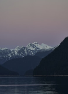Snow-capped mountains rise in the background under a calming gradient sky transitioning from soft pink to blue. A serene body of water reflects the surrounding landscape, bordered by dense, dark forests.