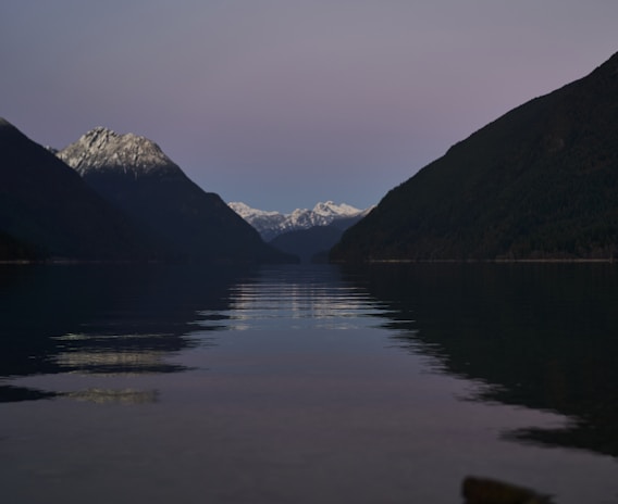 A serene, dark lake reflecting the outline of towering mountains at dusk.