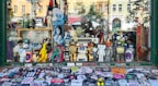 A shop window display filled with various action figures and collectibles. The display includes characters from comics, movies, and video games, such as superheroes, robots, and cartoon icons. The backdrop features a variety of posters and a skateboard, while the window sill is covered with numerous colorful stickers.