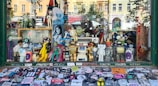 A shop window display filled with various action figures and collectibles. The display includes characters from comics, movies, and video games, such as superheroes, robots, and cartoon icons. The backdrop features a variety of posters and a skateboard, while the window sill is covered with numerous colorful stickers.