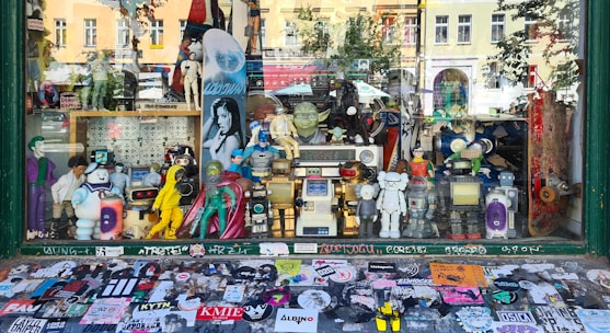 A shop window display filled with various action figures and collectibles. The display includes characters from comics, movies, and video games, such as superheroes, robots, and cartoon icons. The backdrop features a variety of posters and a skateboard, while the window sill is covered with numerous colorful stickers.