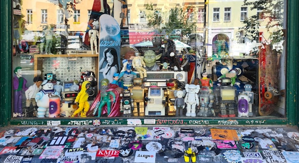 A shop window display filled with various action figures and collectibles. The display includes characters from comics, movies, and video games, such as superheroes, robots, and cartoon icons. The backdrop features a variety of posters and a skateboard, while the window sill is covered with numerous colorful stickers.