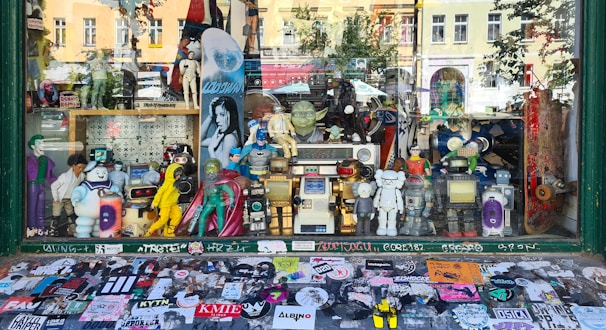 A shop window display filled with various action figures and collectibles. The display includes characters from comics, movies, and video games, such as superheroes, robots, and cartoon icons. The backdrop features a variety of posters and a skateboard, while the window sill is covered with numerous colorful stickers.