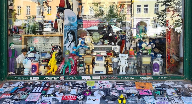 A shop window display filled with various action figures and collectibles. The display includes characters from comics, movies, and video games, such as superheroes, robots, and cartoon icons. The backdrop features a variety of posters and a skateboard, while the window sill is covered with numerous colorful stickers.