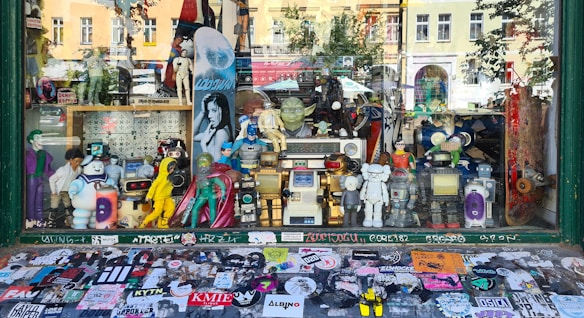A shop window display filled with various action figures and collectibles. The display includes characters from comics, movies, and video games, such as superheroes, robots, and cartoon icons. The backdrop features a variety of posters and a skateboard, while the window sill is covered with numerous colorful stickers.