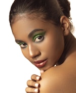 A close-up portrait of a person with vibrant green eyeshadow and metallic lipstick, looking over their shoulder with a serene expression. The focus is on their face and makeup, while the background is plain, drawing attention to the subject.