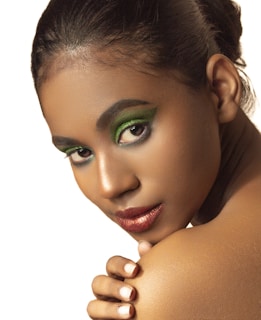 A close-up portrait of a person with vibrant green eyeshadow and metallic lipstick, looking over their shoulder with a serene expression. The focus is on their face and makeup, while the background is plain, drawing attention to the subject.